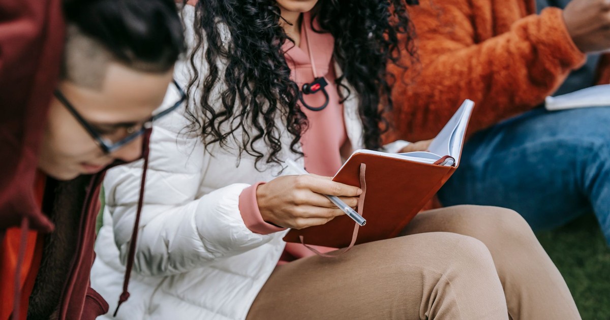 Crop unrecognizable multiracial classmates with paper notepads sitting on grass and studying together