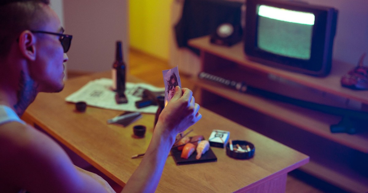 A tattooed man examines a photo in a dimly lit room with a retro TV.