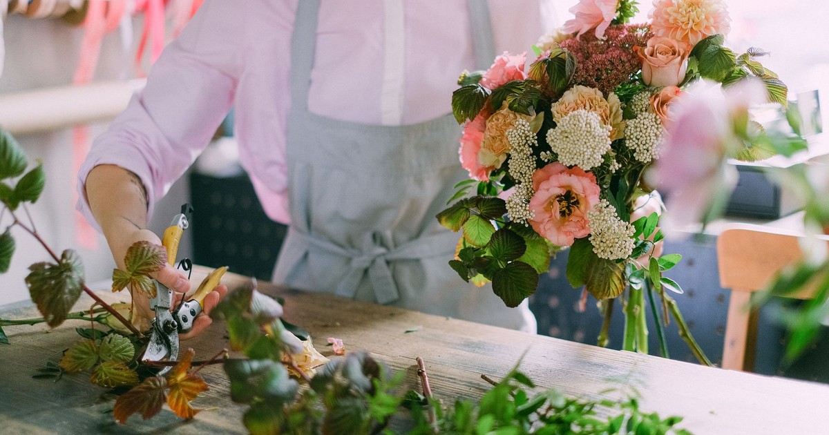 A florist wearing an apron and long sleeves is arranging a vibrant flower bouquet with pruning shears indoors.