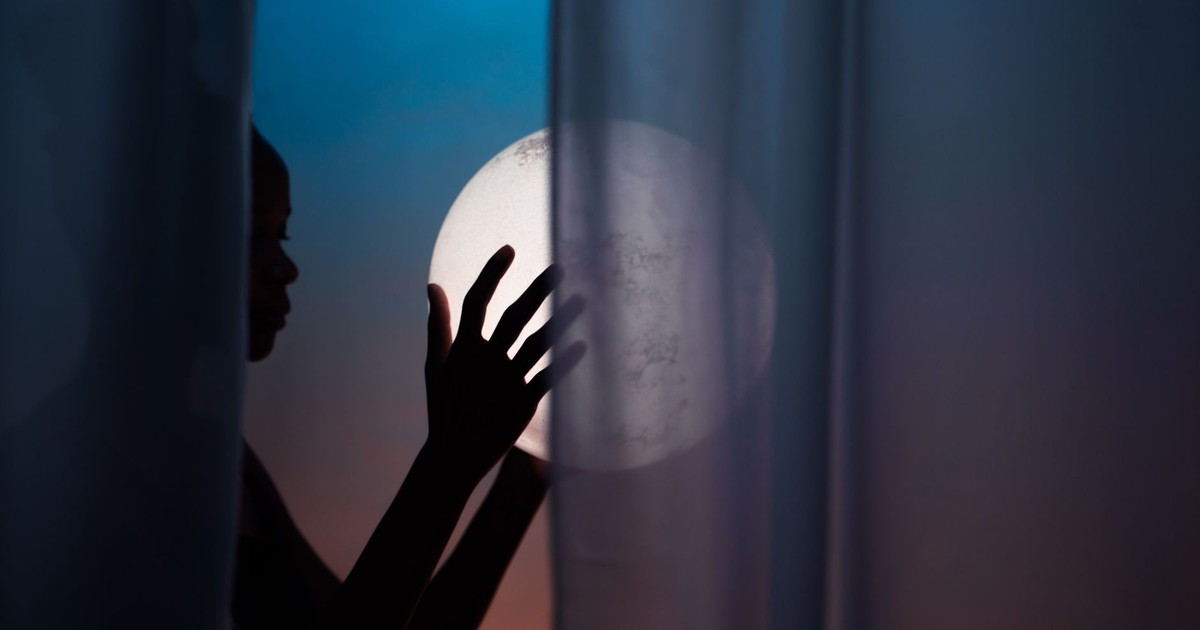 Artistic silhouette of a person holding a glowing orb behind sheer curtains at twilight.