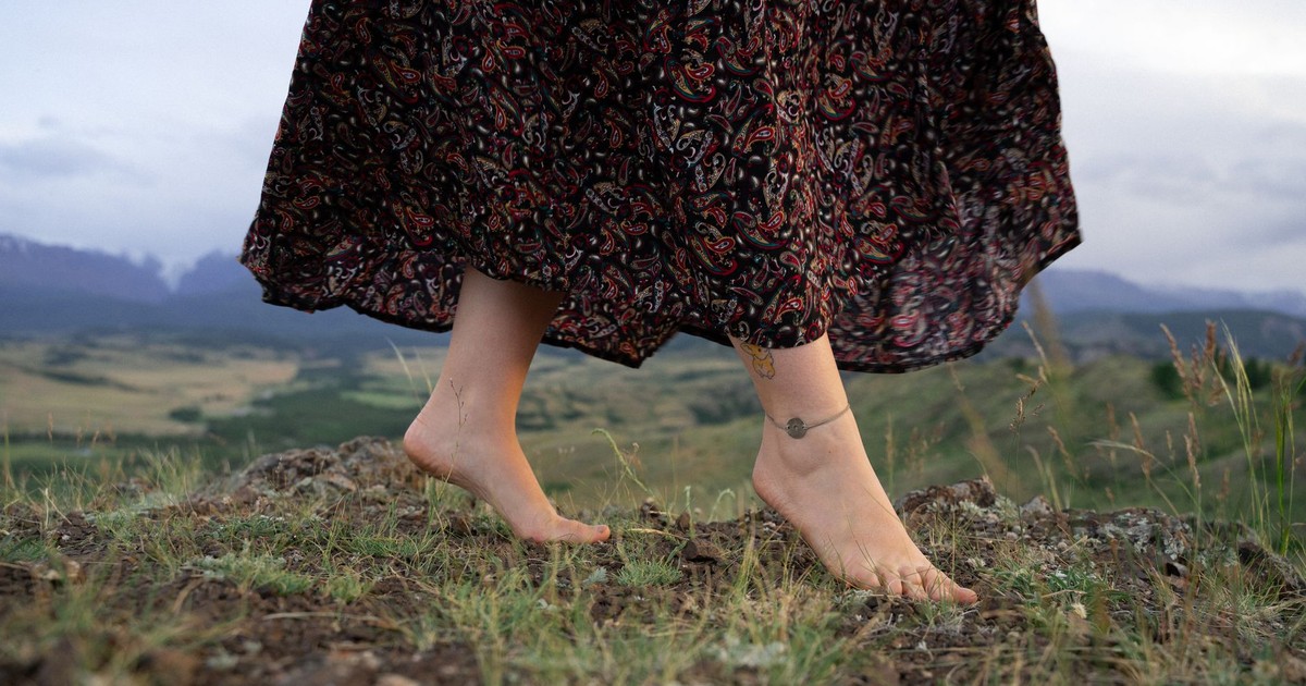 Close-up of a woman walking barefoot on grassy terrain, showcasing anklet and patterned skirt in a natural setting.