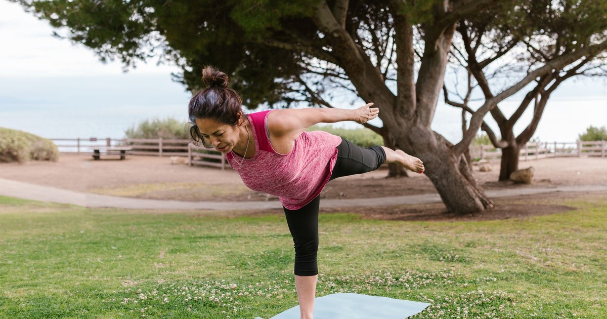 Woman in a balancing yoga pose on a mat in a scenic outdoor park setting.