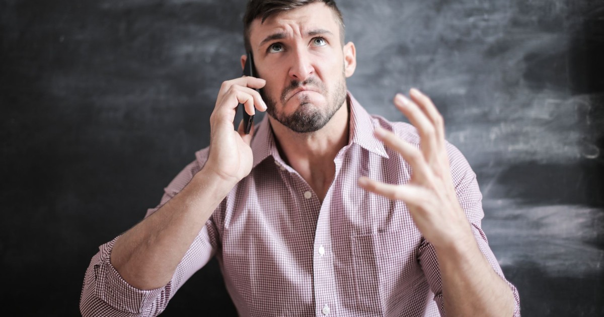 Adult man in pink dress shirt looking stressed while talking on a smartphone indoors.