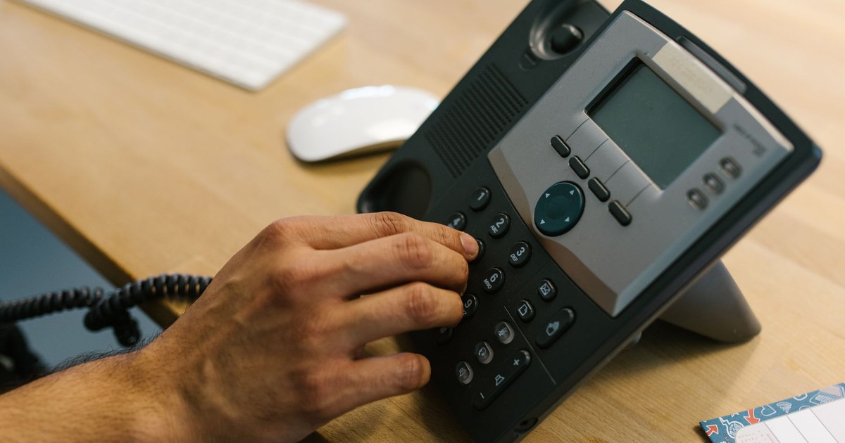 A hand dialing a phone on a wooden desk. Office environment with keyboard and planner.