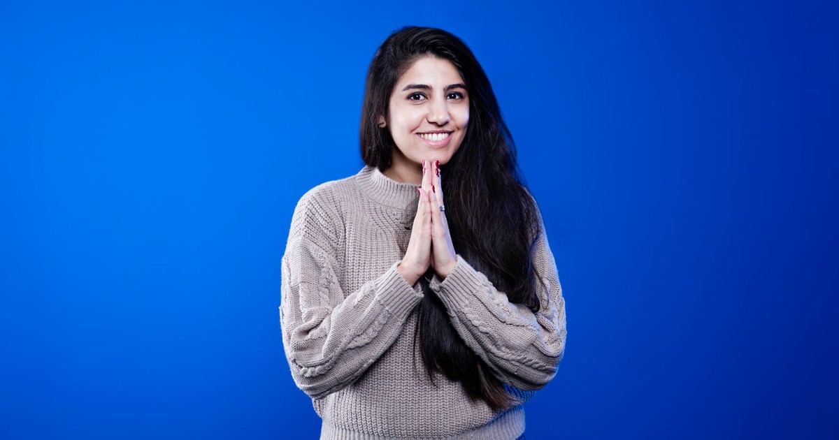 Happy woman with long hair wearing a sweater, smiling with hands in prayer position against a blue background.