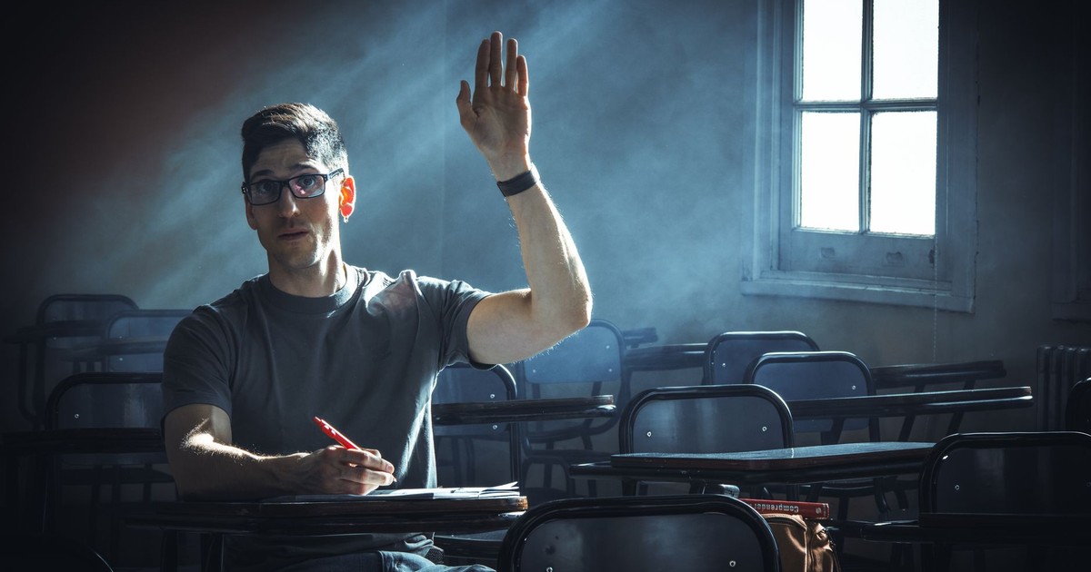 Person sitting in a dimly lit classroom raising hand, conveying attention and inquiry.