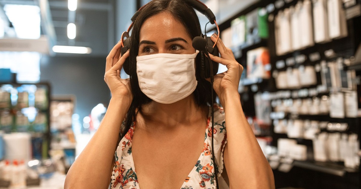A woman in a store tries on headphones while wearing a face mask, showcasing modern shopping.