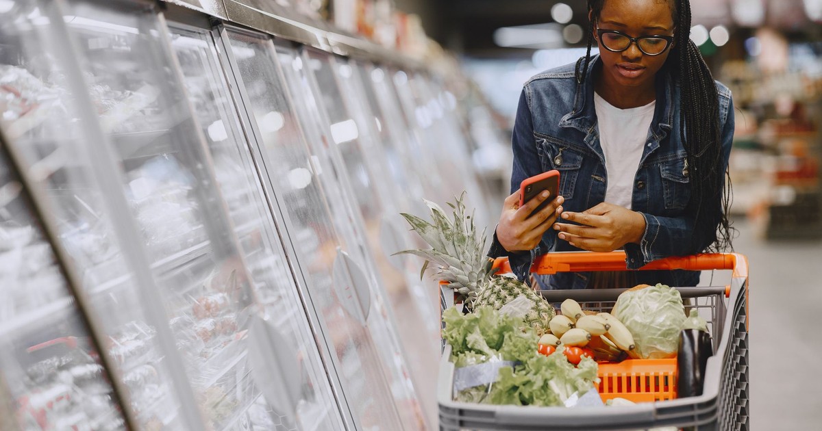Woman in a grocery store using smartphone while pushing a shopping cart filled with groceries.