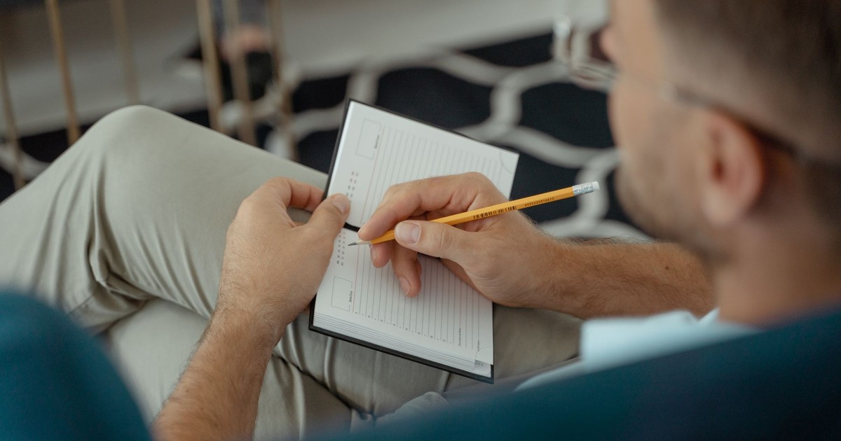 An adult man writing with a pencil in a notebook while seated indoors.