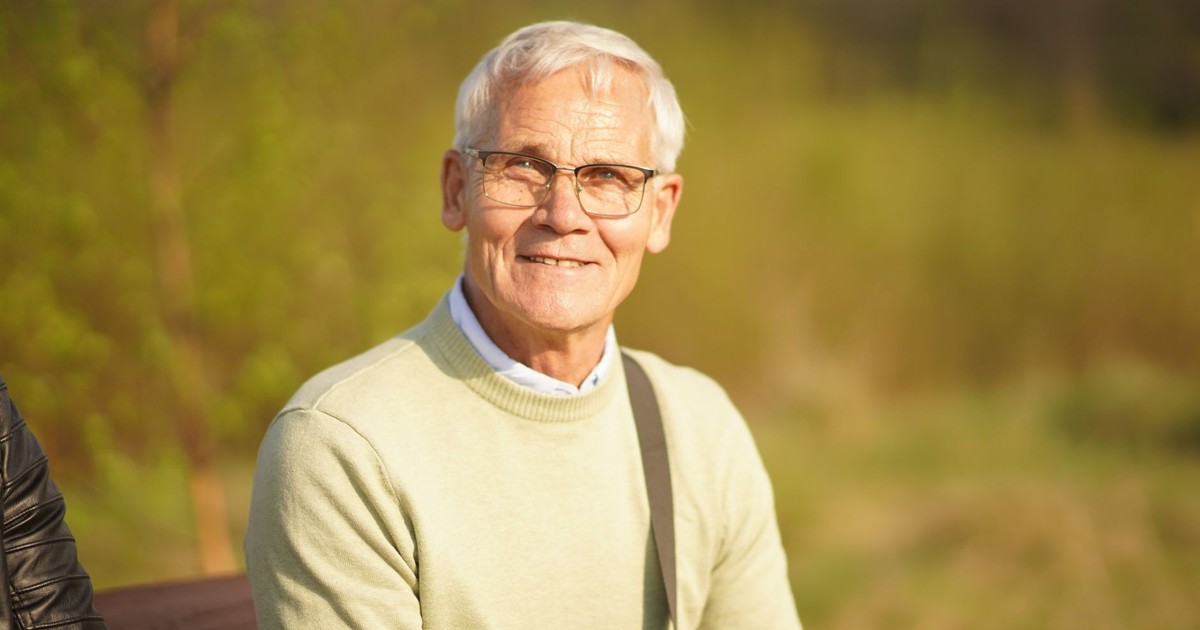 Senior man smiling warmly while sitting outdoors in bright sunlight, wearing glasses.