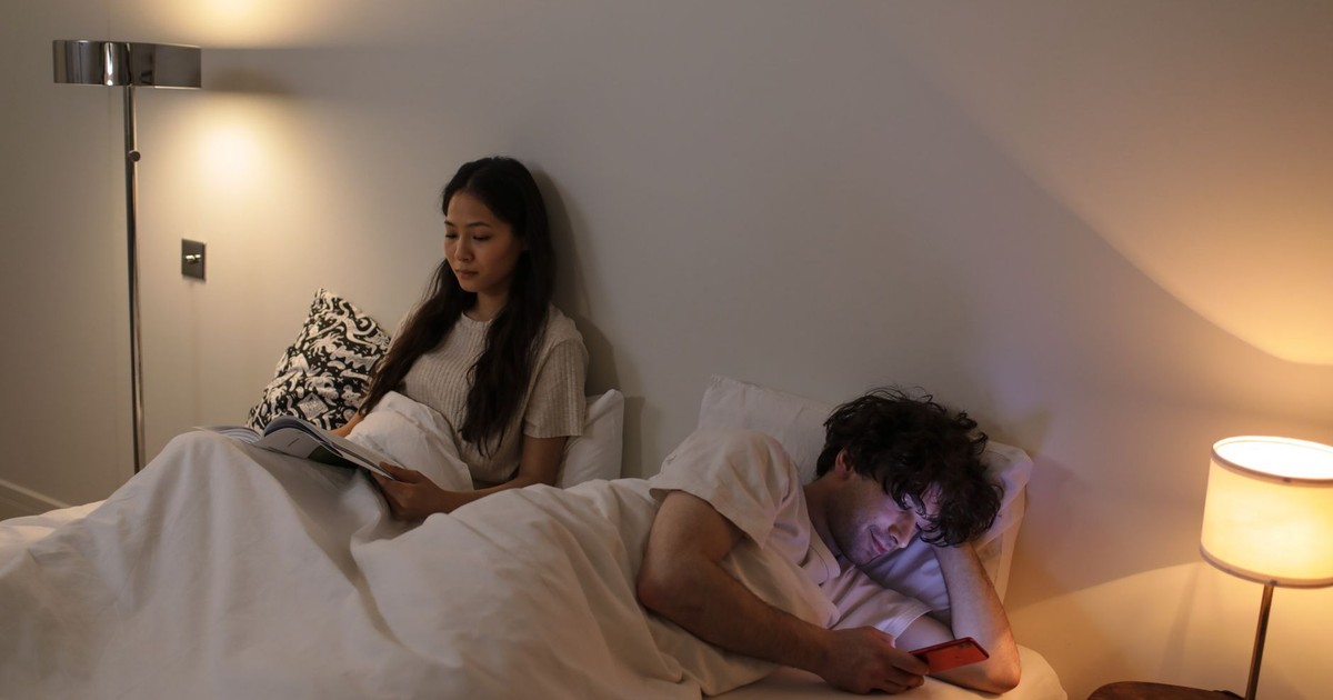 A couple in bed, one reading and the other using a phone, with warm lighting.