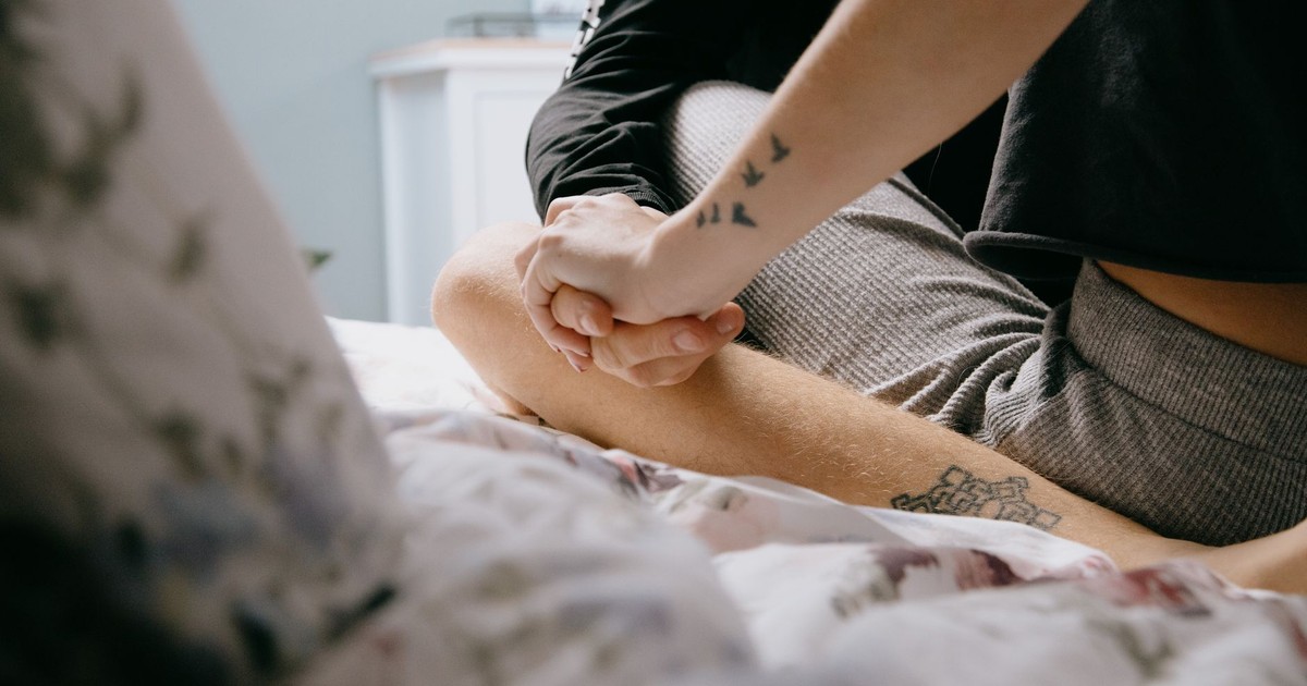 Crop faceless couple with stylish tattoos sitting on bed with crossed legs and holding hands