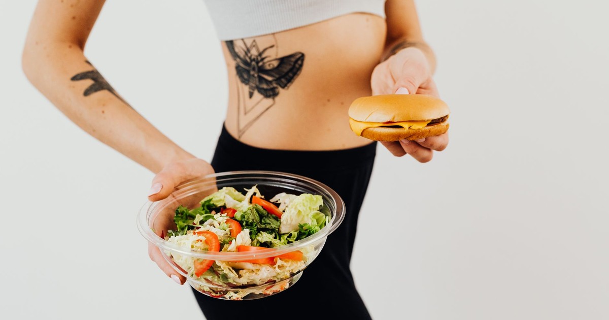 A woman with a tattoo holding a healthy vegetable salad and a cheeseburger, representing food choices.