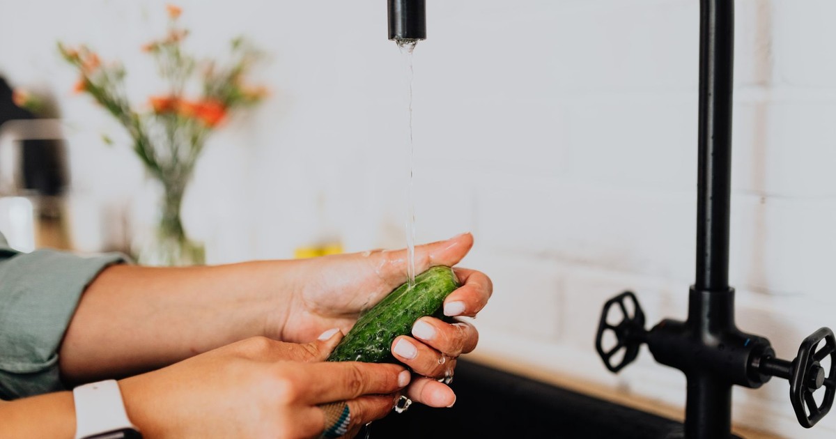 Hands washing a fresh cucumber under running water in a kitchen setting.