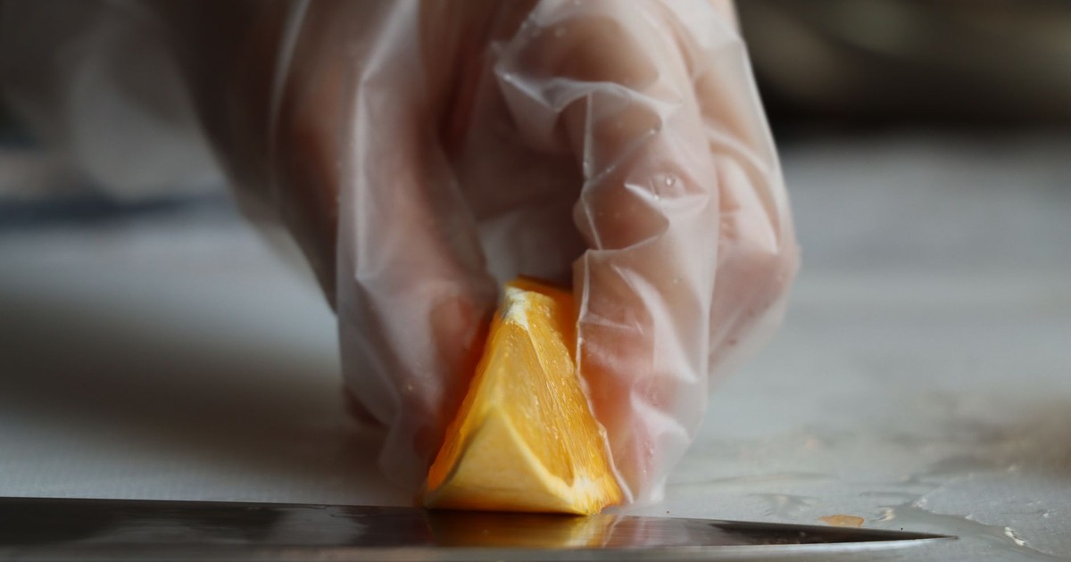 Gloved hand carefully slices an orange wedge with a knife, showcasing precise cutting technique.