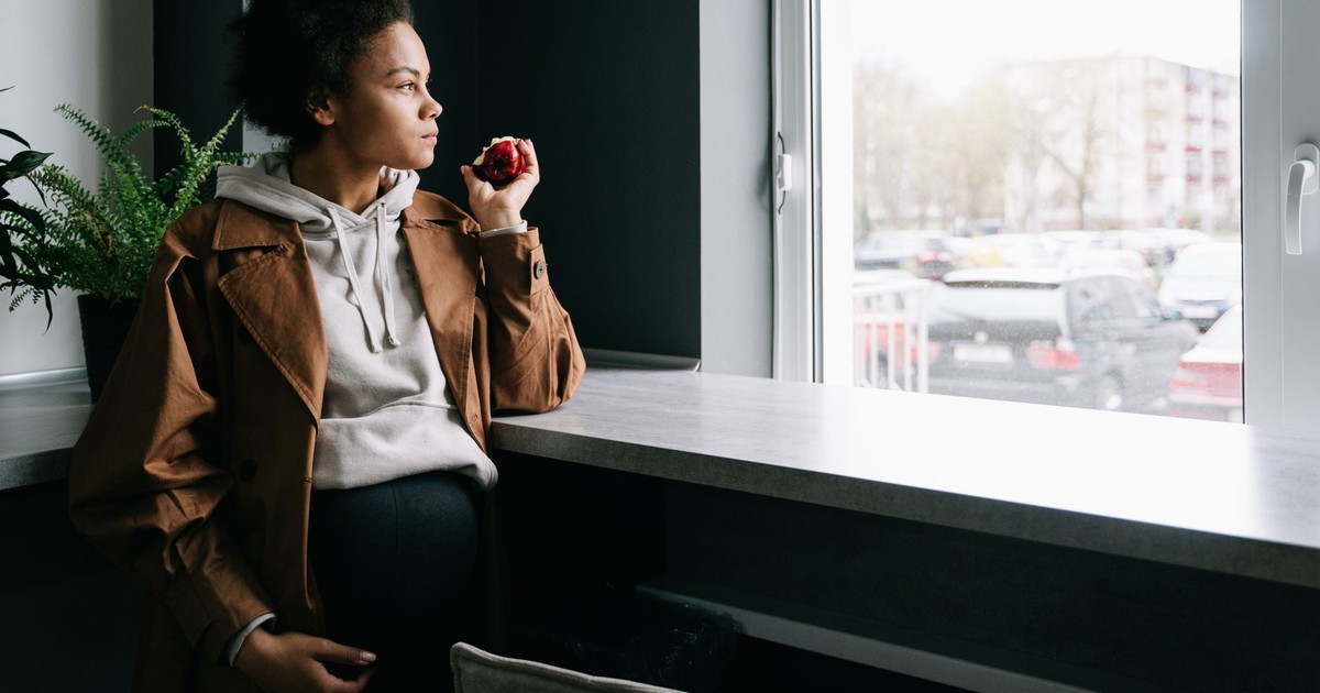 Pregnant woman in casual attire holding an apple, gazing thoughtfully out a window.