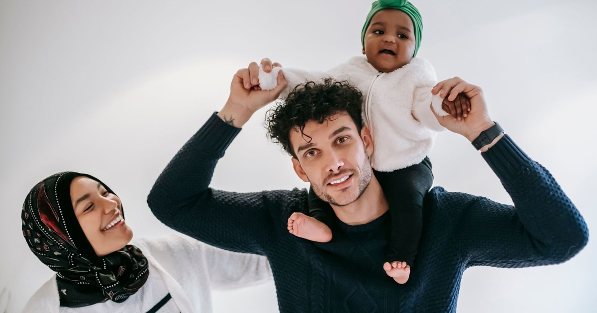 Positive dad enjoying fatherhood with girl on shoulders near Muslim mother in traditional hijab against white background