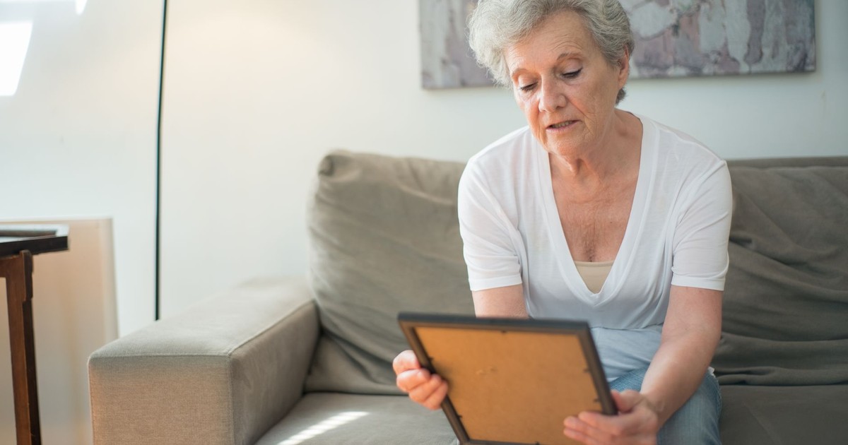 Senior woman sitting indoors, holding a picture frame and reminiscing, evokes feelings of nostalgia and contemplation.