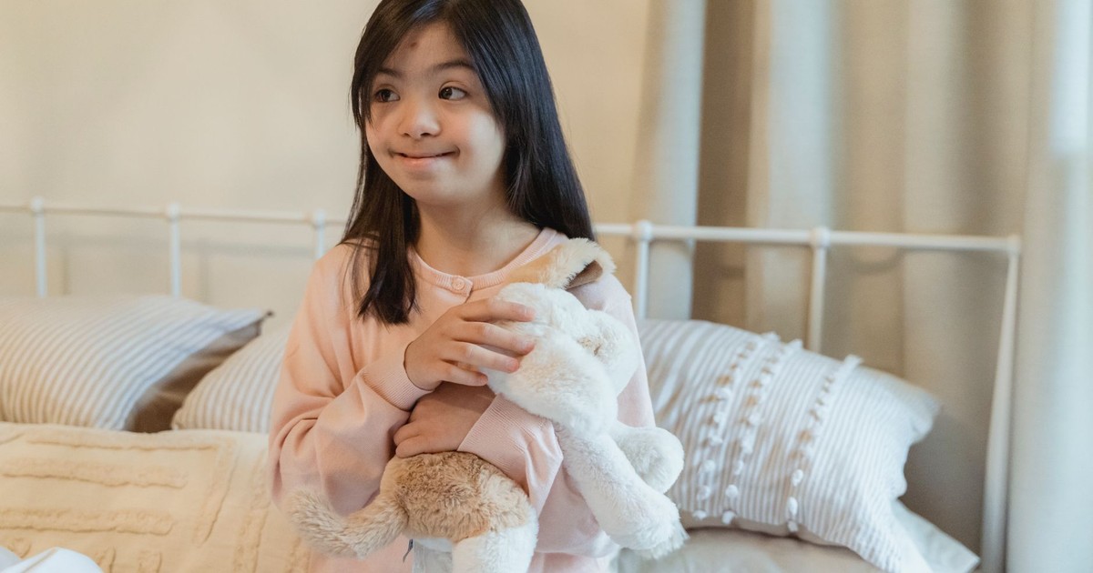 A smiling young girl in a bedroom holding a toy rabbit while sitting on a bed.