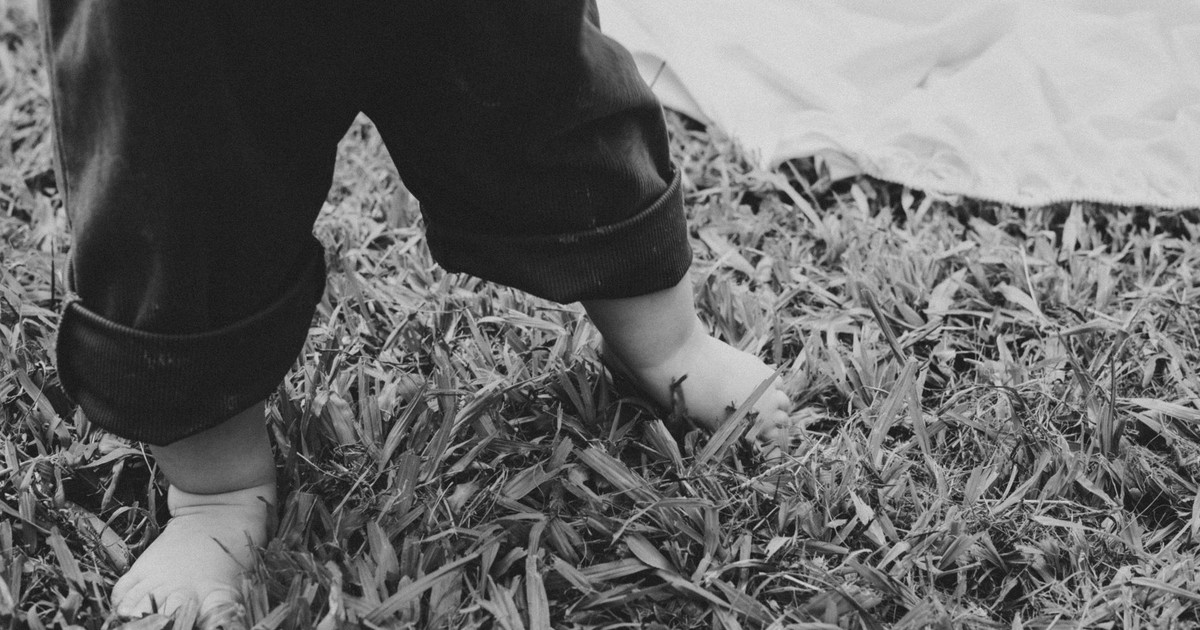 Black and white photo of a baby taking first steps on grass, showing feet and rolled-up pants.