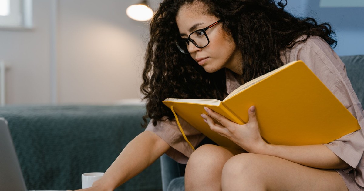 Woman with curly hair studying indoors, using a laptop and taking notes in a large notebook.