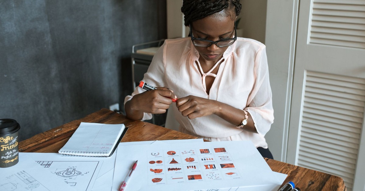 African woman with dreadlocks working on diagrams at an office desk, wearing eyeglasses and a blouse.