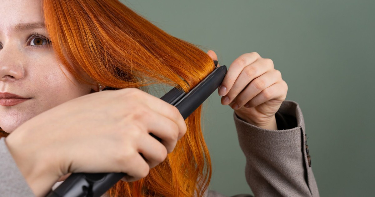 Close-up of a woman styling her red hair using a hair straightener against a green background.