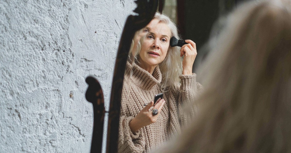 Elderly woman puts on makeup while looking into a mirror at home.