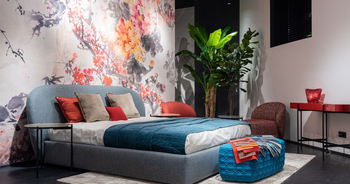 Comfortable bed with colorful cushions and blue blanket placed at painted wall in modern bedroom with armchairs and potted plant