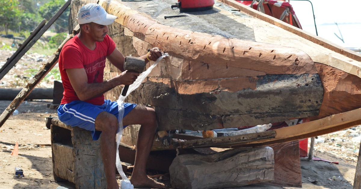 A man works with a mallet to repair a damaged wooden boat in a sunny outdoor setting.