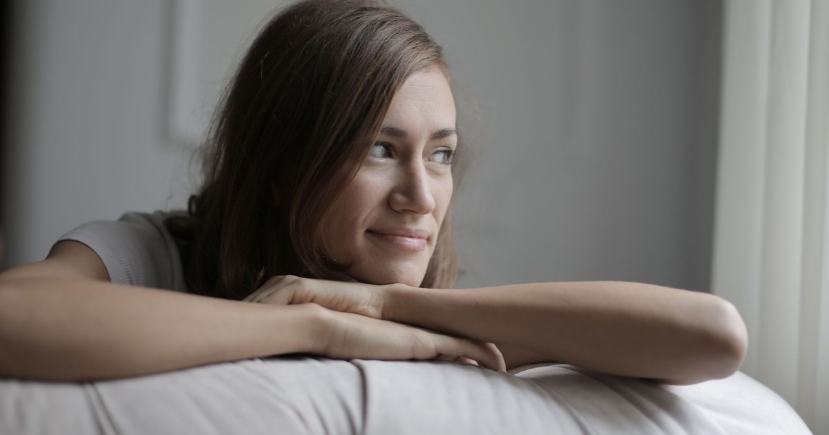 A young woman rests on a sofa inside, smiling gently while looking out the window.