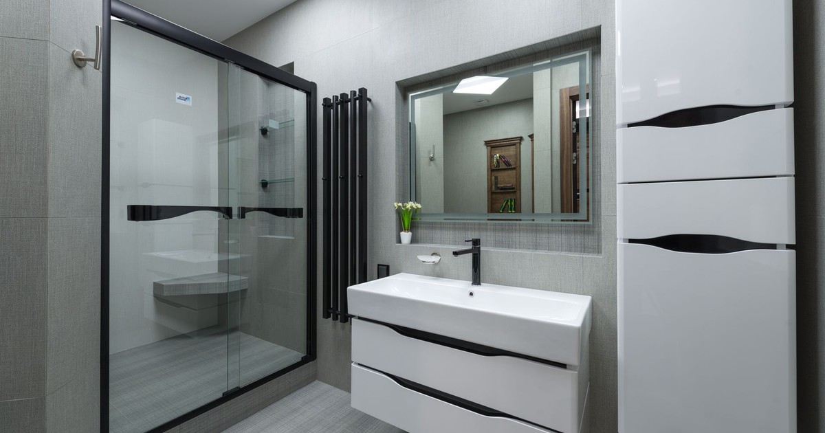 White sink at wall with mirror near cupboard in modern bathroom with glass shower cabin and bright illumination in apartment
