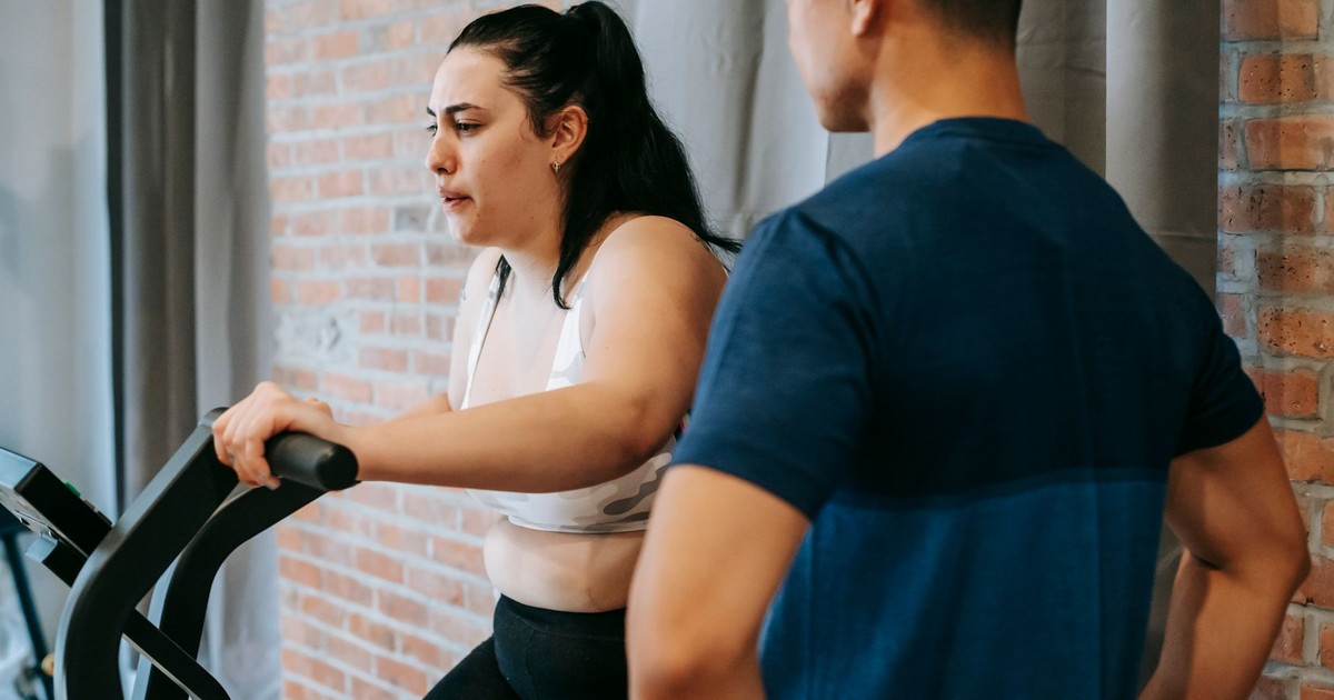 Side view of serious concentrated young obese female doing weight lose exercise on cycling equipment during cardio training with crop ethnic male personal coach