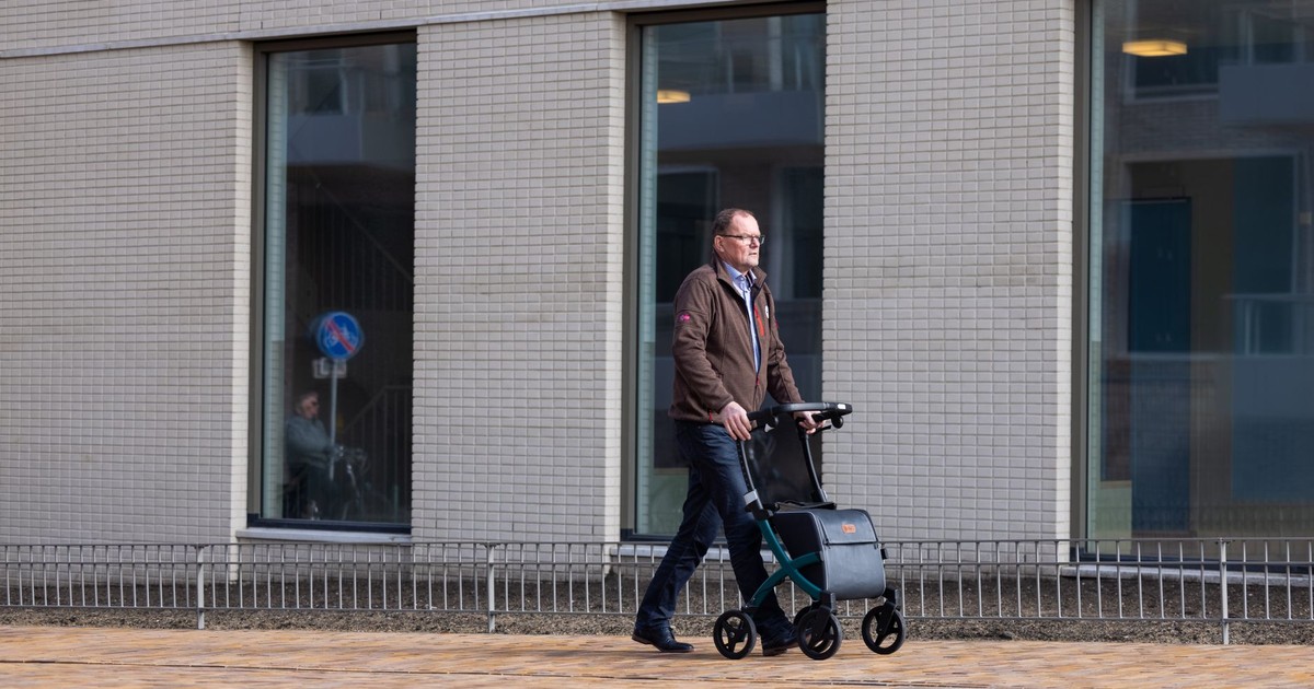 Elderly man using a rollator for support while walking outside a building in an urban area.