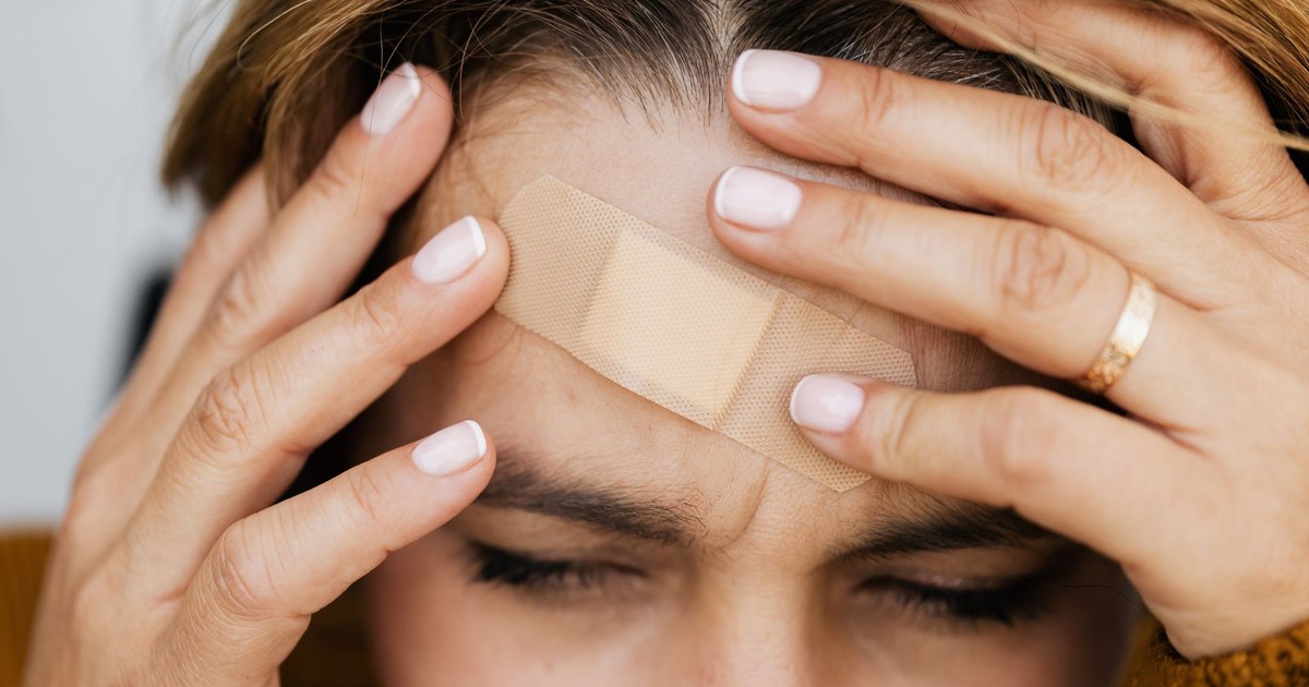 A person with a band aid on their forehead holds their head in pain, eyes closed.