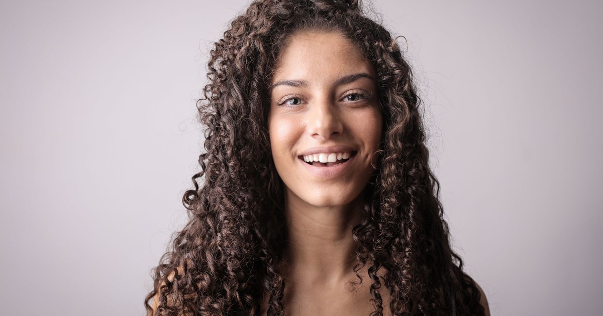 A cheerful woman with curly hair smiles warmly against a gray background.