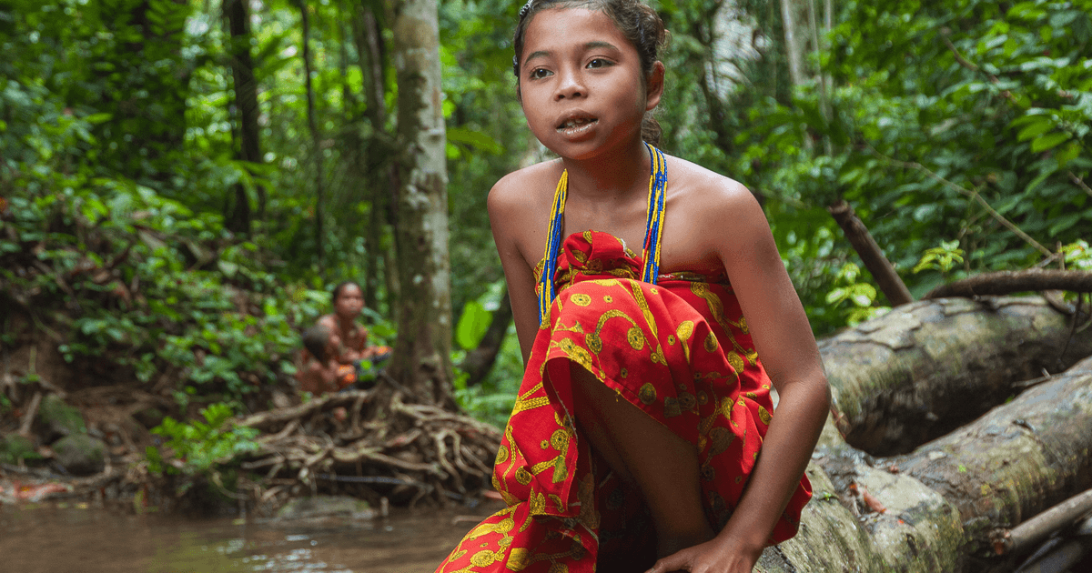 A young girl in traditional attire sits on a log in a vibrant jungle setting.