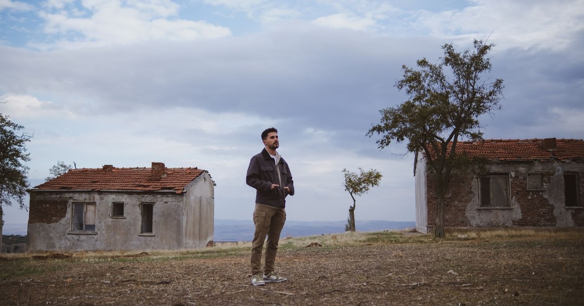 A man stands in front of old, abandoned houses in a rural area with a cloudy sky.