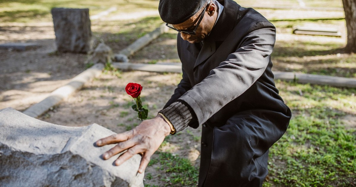 A senior adult kneeling by a gravestone holding a red rose, symbolizing mourning in a cemetery.