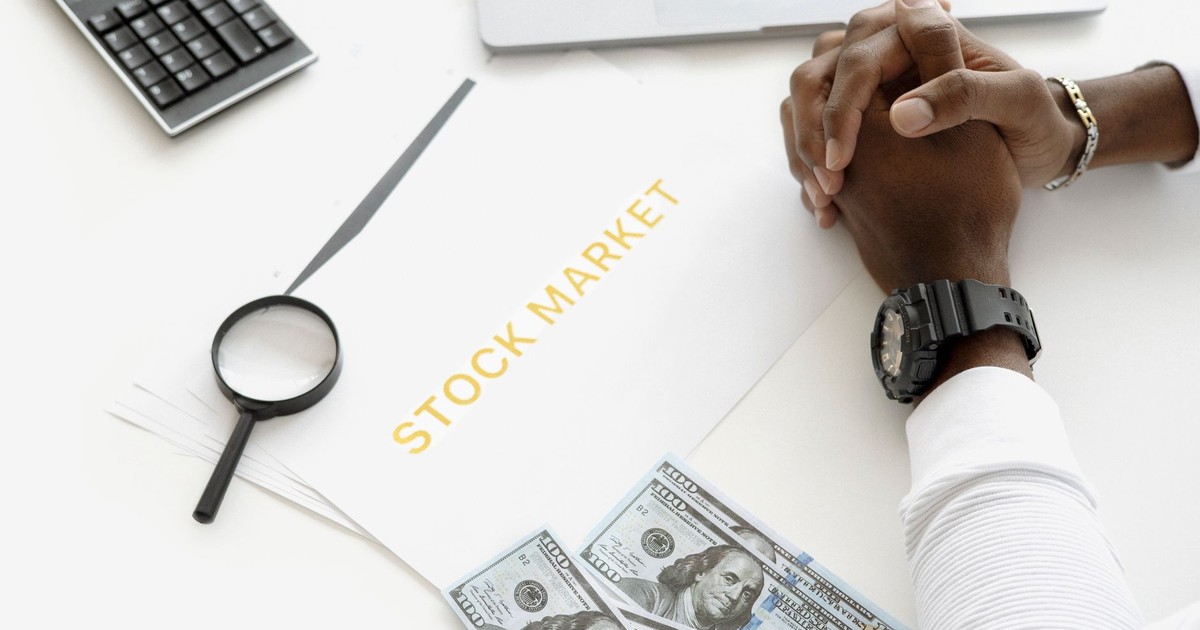 Hands folded over stock market documents with calculator and cash on desk.