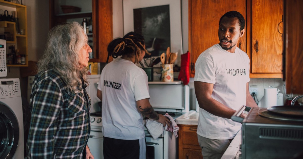 Volunteers interact and assist a senior in a cozy kitchen environment, promoting community support.