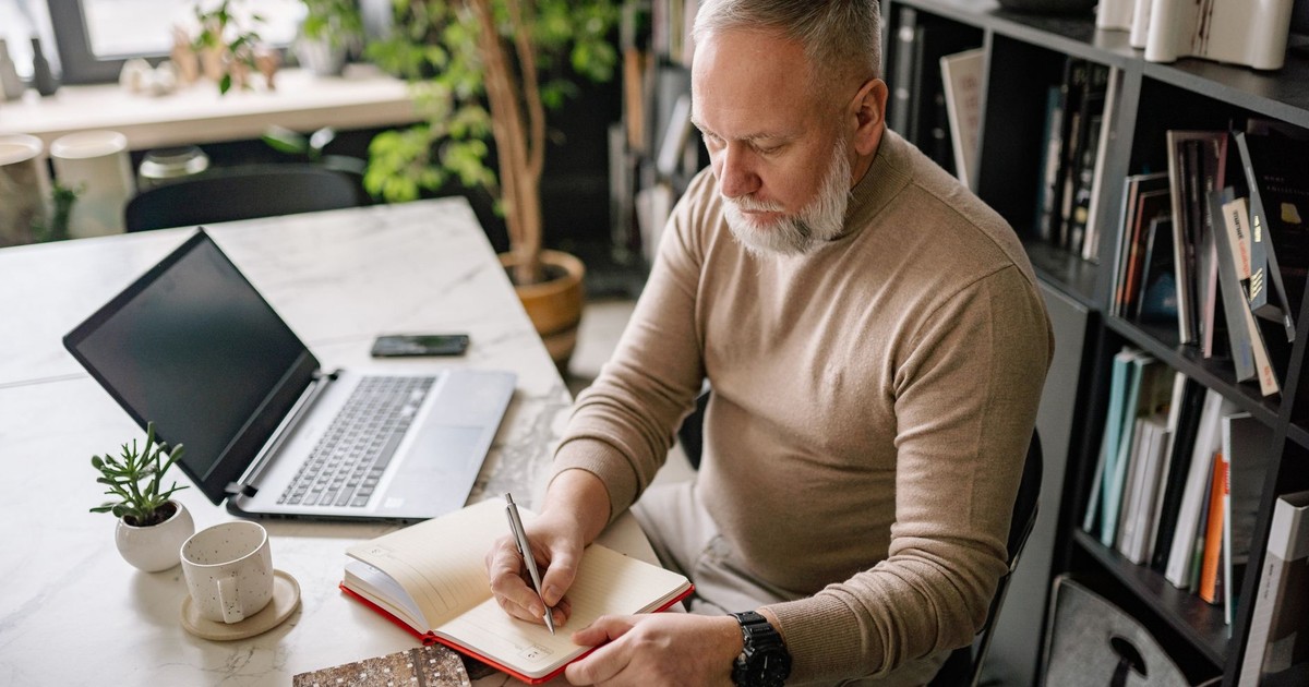 A mature man with a beard writes in a diary at his home office desk next to a laptop.