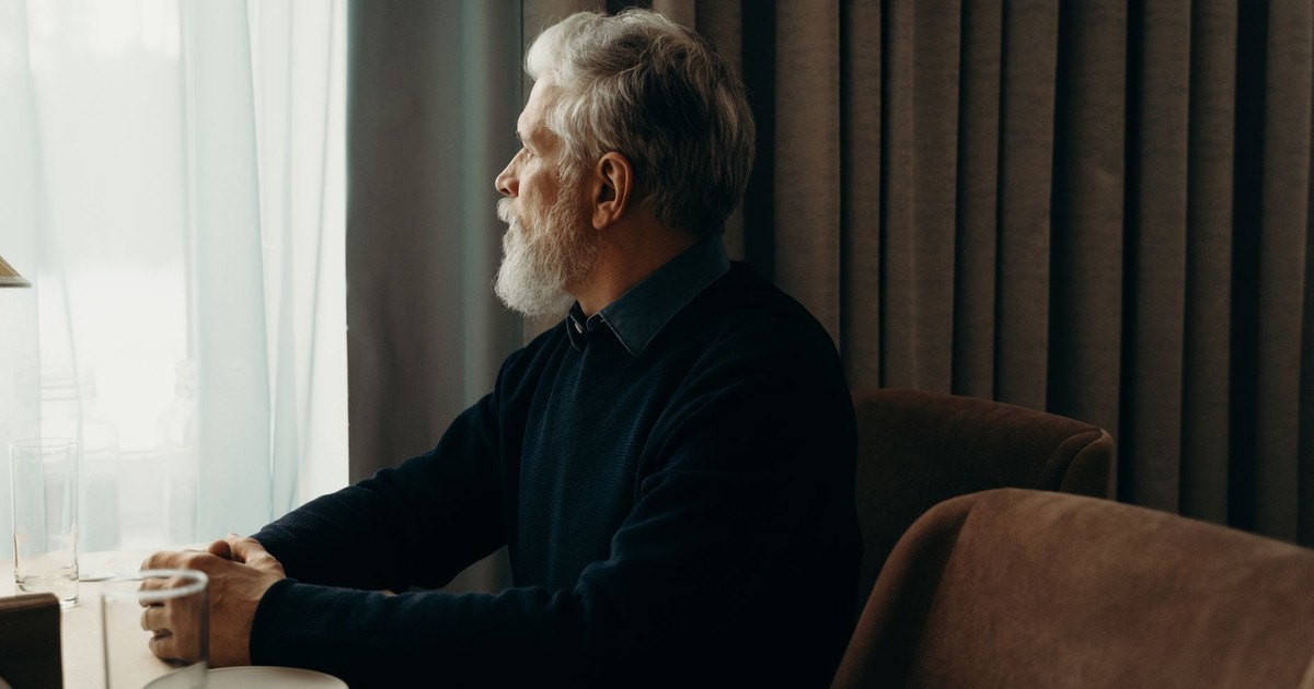 An elderly man with a beard looking outside while sitting by a table indoors.
