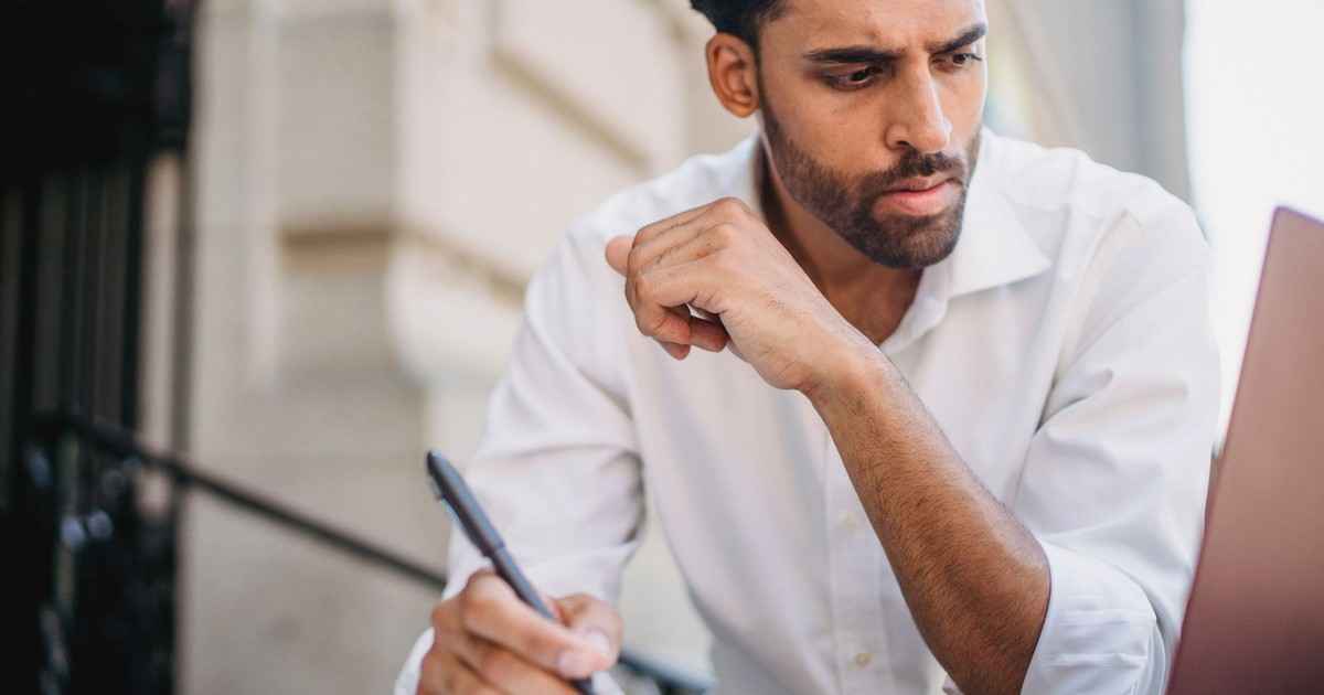Focused man in white shirt takes notes while working remotely on his laptop outside, exuding concentration.