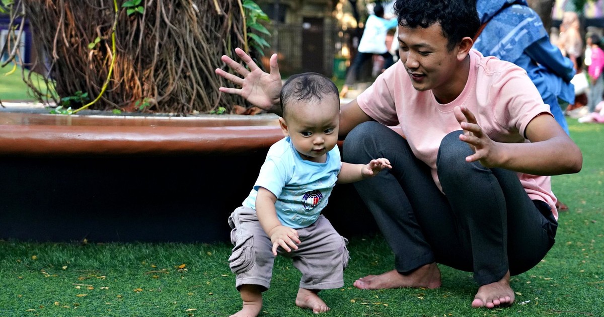 A father encourages his baby to take first steps on a sunny day in a park, barefoot on green grass.