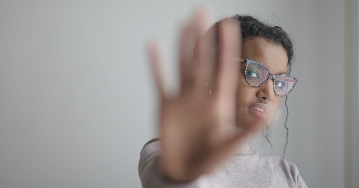 Young African American female wearing casual clothes and eyeglasses outstretching arm at camera while standing near grey wall