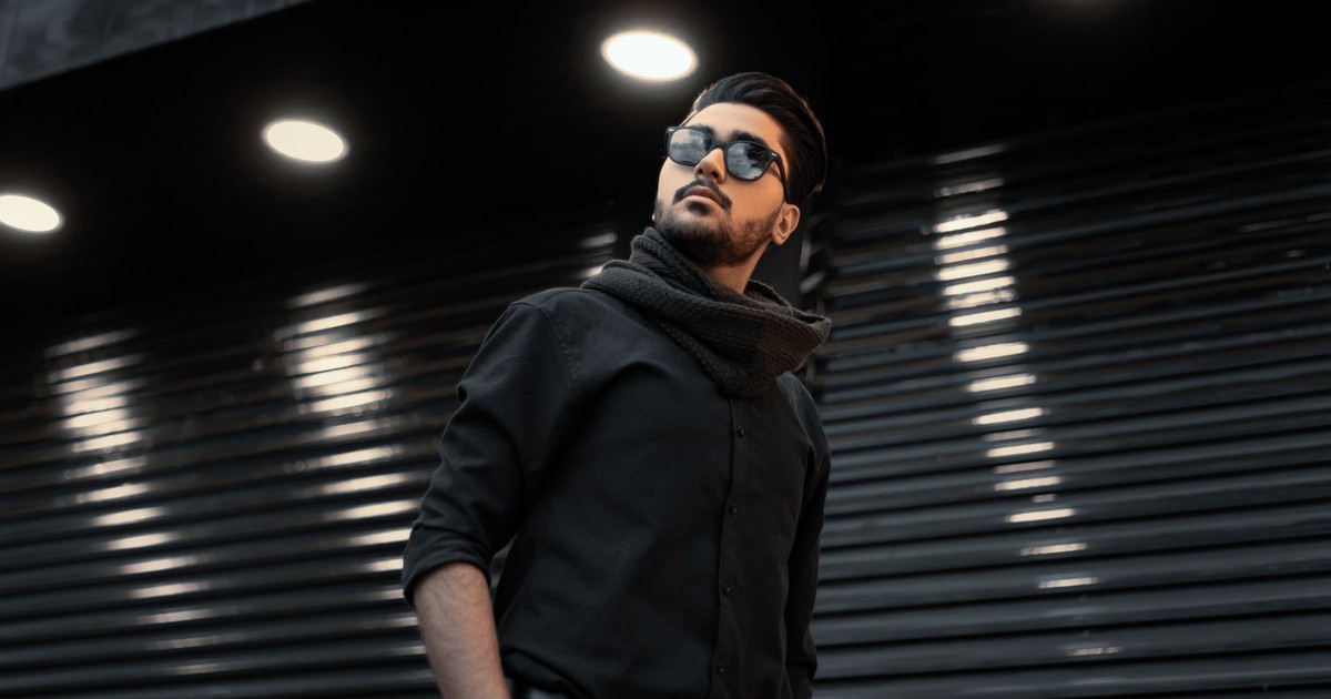 A fashionable man in sunglasses with a scarf poses under spotlights in an urban setting.