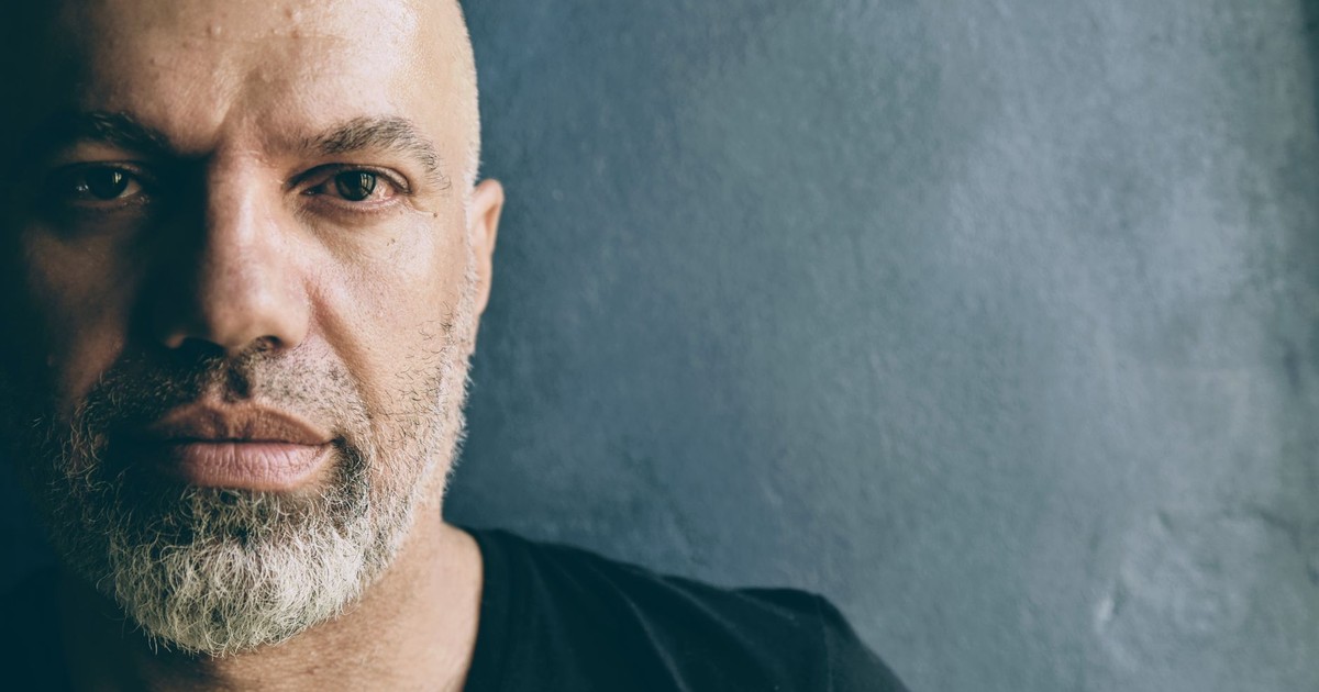 A close-up, moody portrait of a bald man with a beard against a textured background.