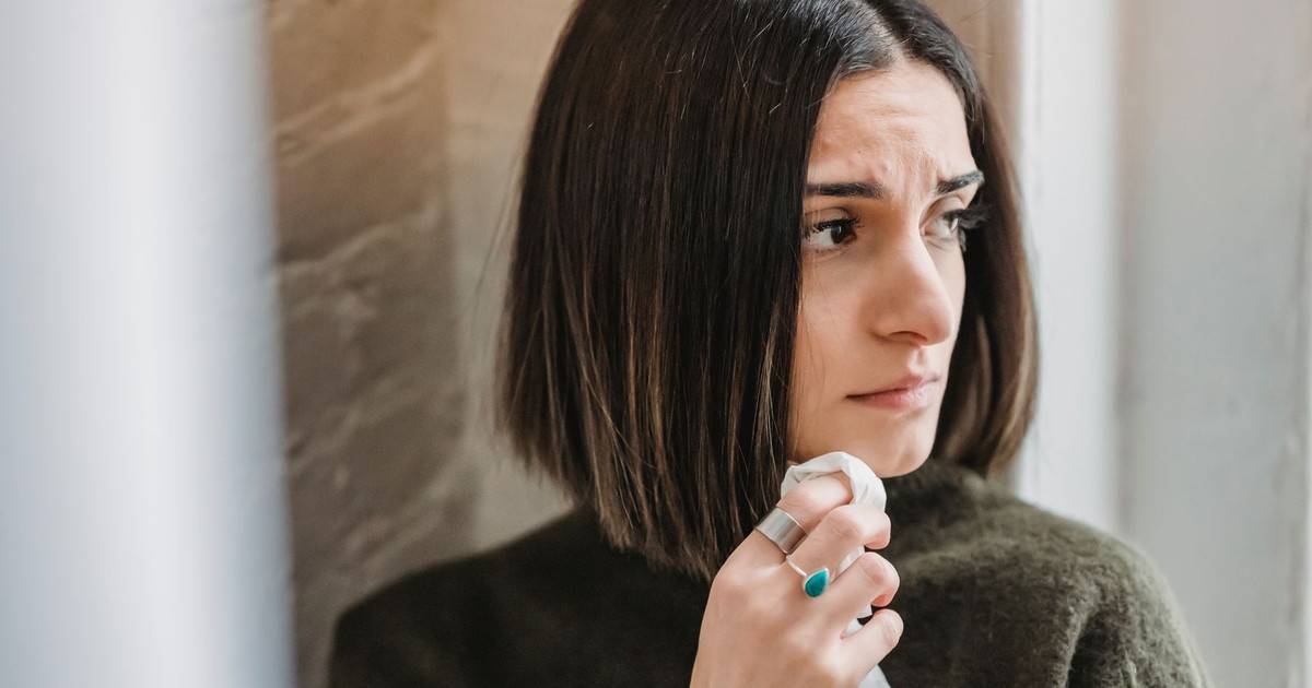 Sad young ethnic female millennial with dark hair standing near brick wall with tissue in hand and looking away thoughtfully