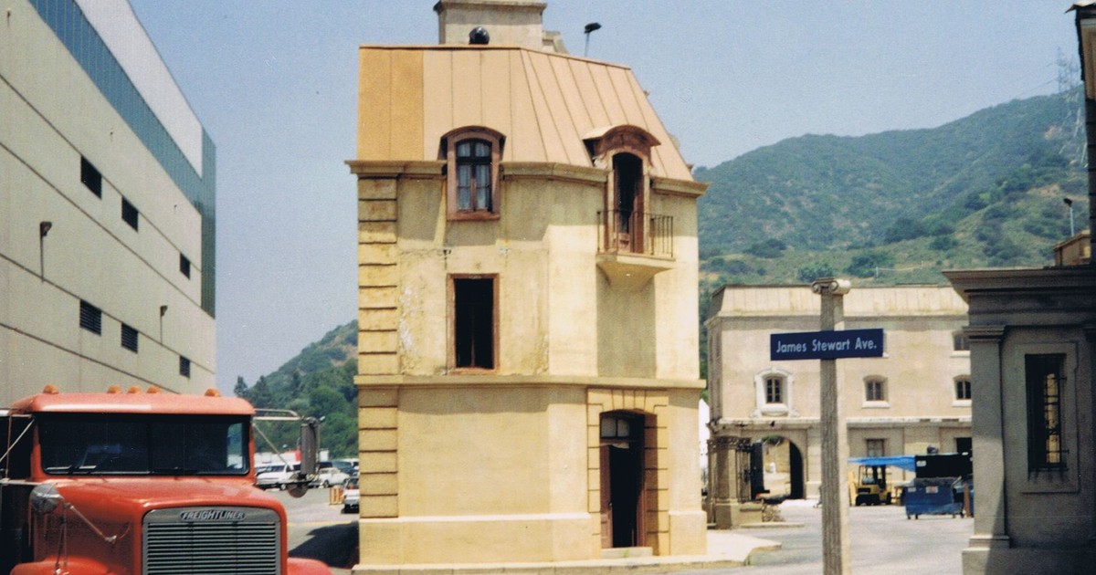 A rustic building on a film studio lot with a parked truck near a mountain background.